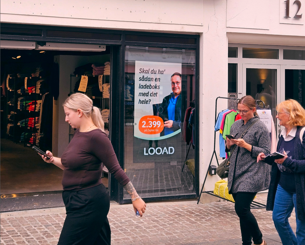 Den digitale EuroSize-skærm i et butiksvindue på Bispensgade 12 i Aalborg med fodgængere, der passerer forbi